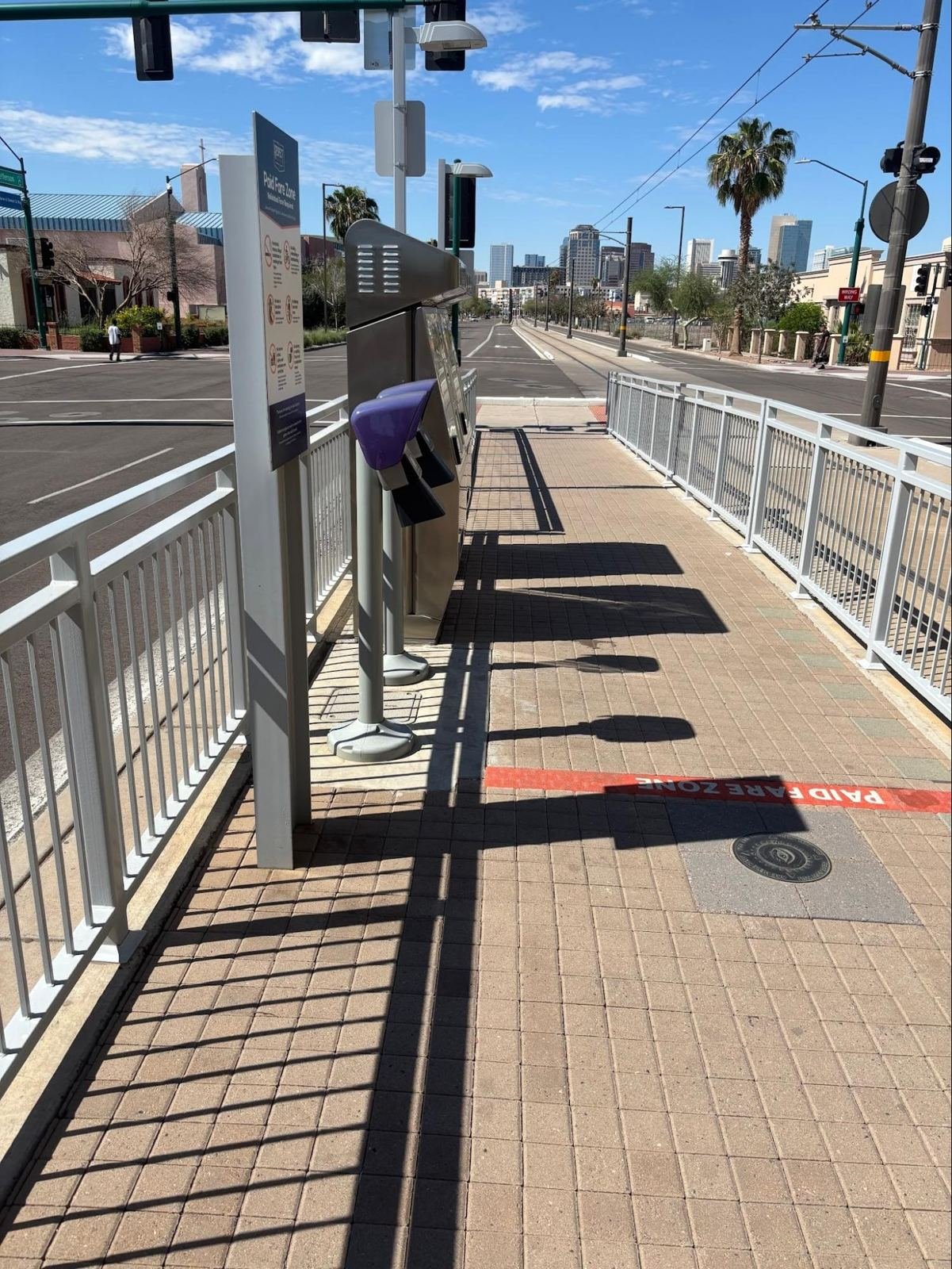Looking down a narrow platform lined with railings: a fare machine and scanners stand in bright sun, casting hard shadows on the pavers.