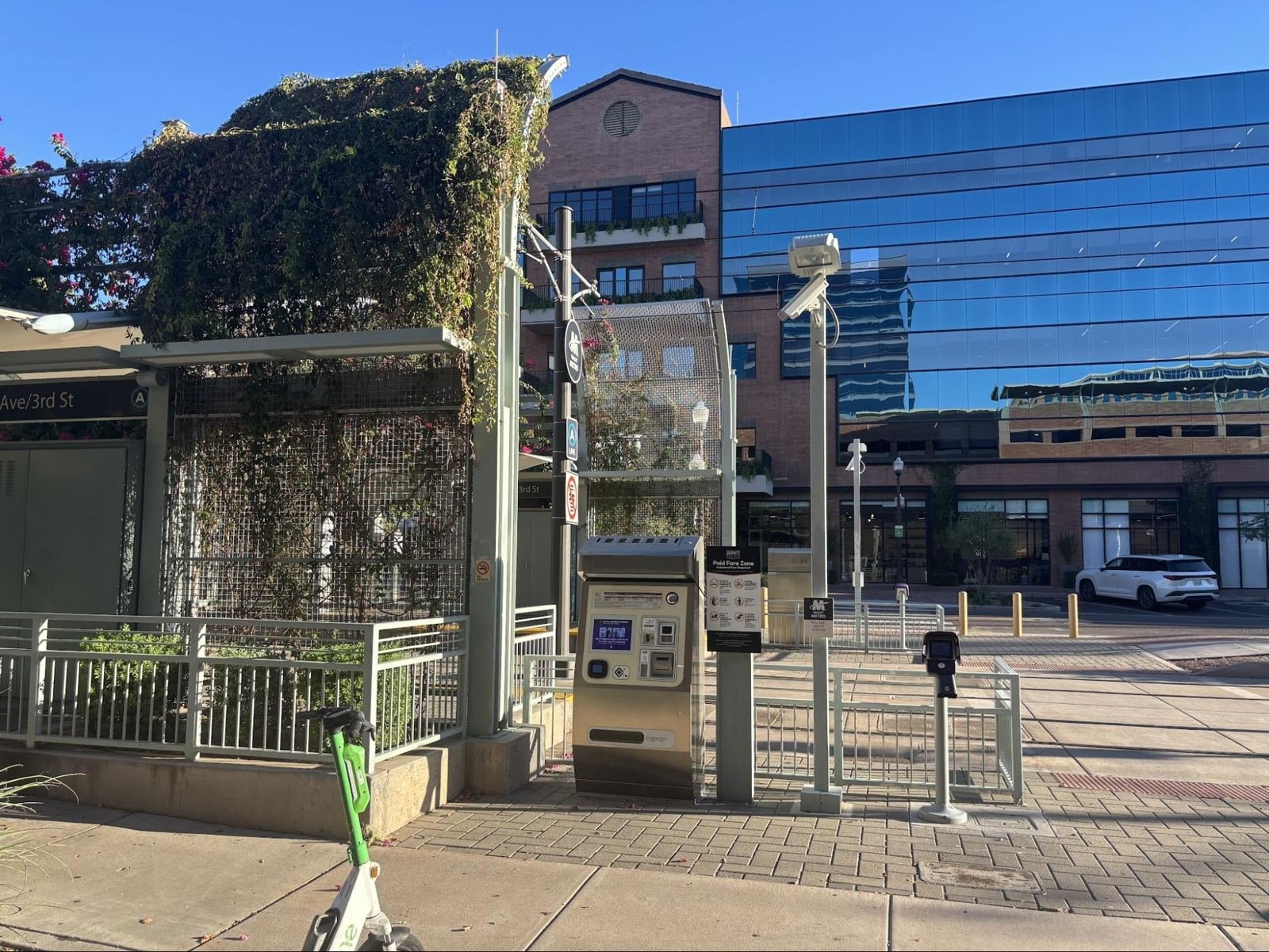 A fare machine and a pass scanner stand in full sun on the sidewalk outside a vine-covered shade structure at the Mill Avenue and 3rd Street station.